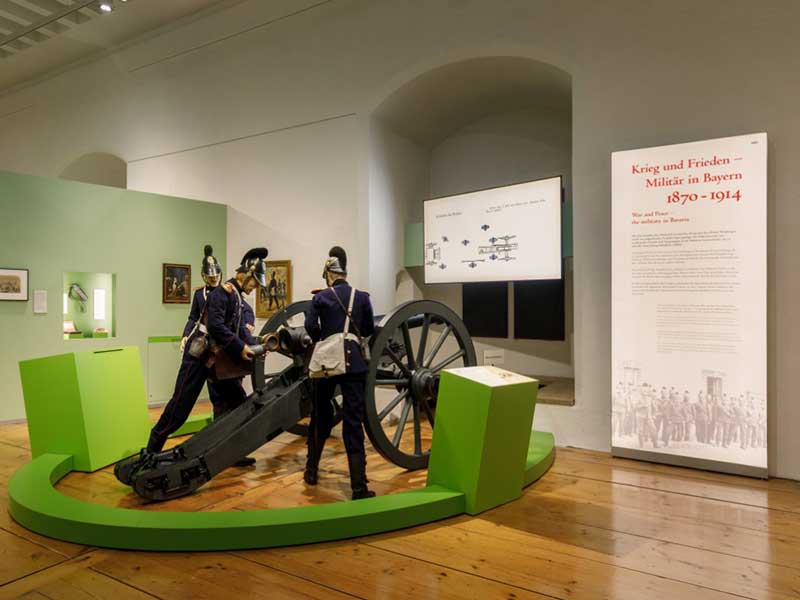 Museumsausstellung mit historischen Soldatenfiguren und einem Geschütz vor Informationstafeln.